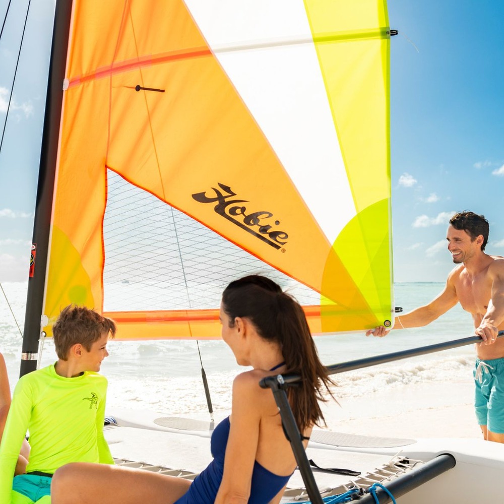 Three people prepare a small sailboat with vivid yellow and orange sails on a sunny beach, setting the scene for an unforgettable destination wedding.