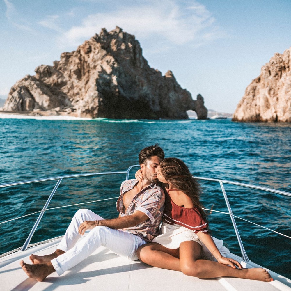 A couple shares a kiss on a boat during their destination wedding, with dramatic rocky cliffs and sparkling blue water providing a breathtaking backdrop.