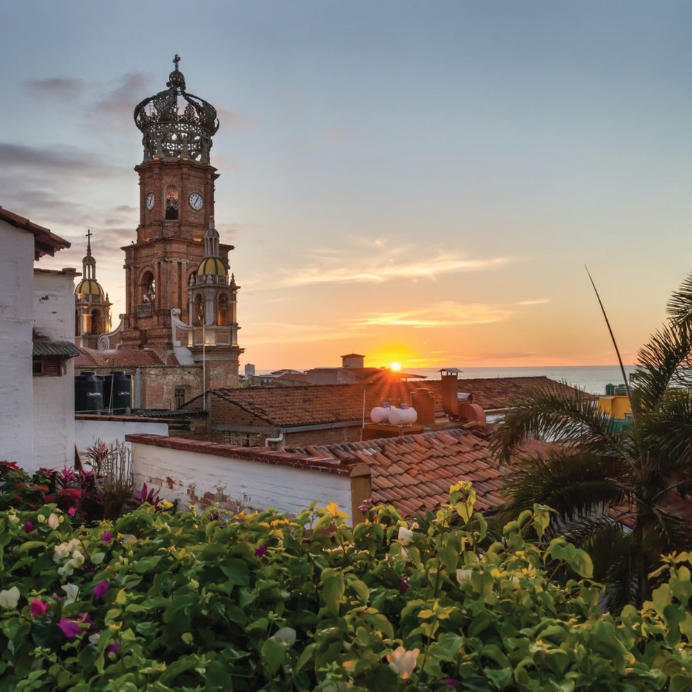 A picturesque church tower rises above the rooftops at sunset, framed by vibrant flowers and swaying palm trees—a dreamy setting perfect for a destination wedding.