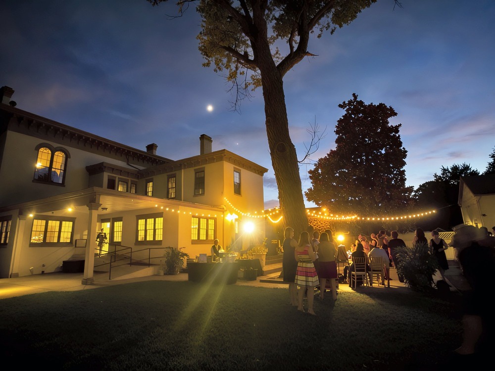 At dusk, guests gather under glowing string lights outside a spacious house with a tree in the yard, celebrating a beautiful destination wedding.