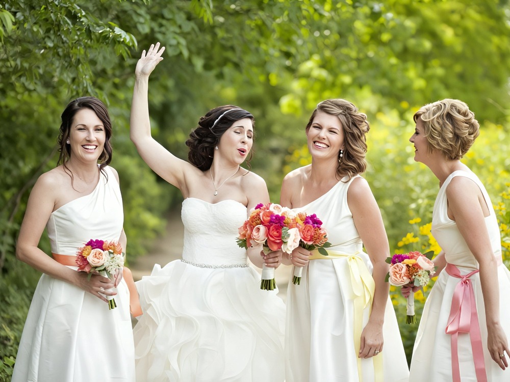 Four women in white dresses, holding bouquets, smile and laugh together outdoors among lush greenery at a destination wedding.