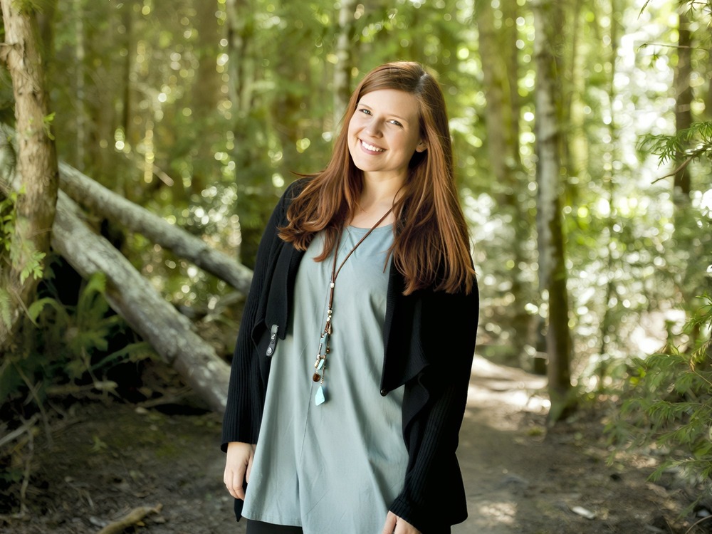 A woman with long brown hair stands on a forest path, smiling at the camera in daylight, capturing a joyful moment during her destination wedding.
