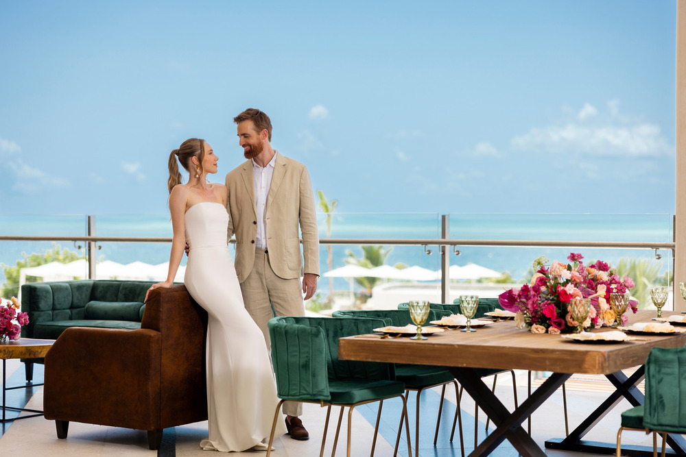 A couple in formal attire stands by a table at a luxury destination wedding, ocean views behind.