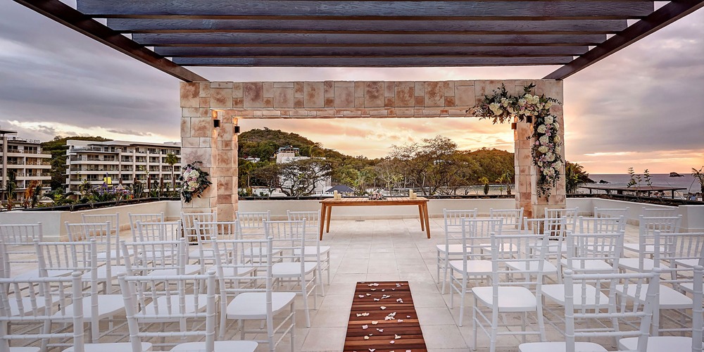 Destination wedding ceremony on a luxury resort rooftop, floral arch and sunset in the background.
