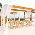 A wooden wedding arch adorned with white drapes stands on the sandy beach, surrounded by chairs arranged for a beautiful destination wedding ceremony.