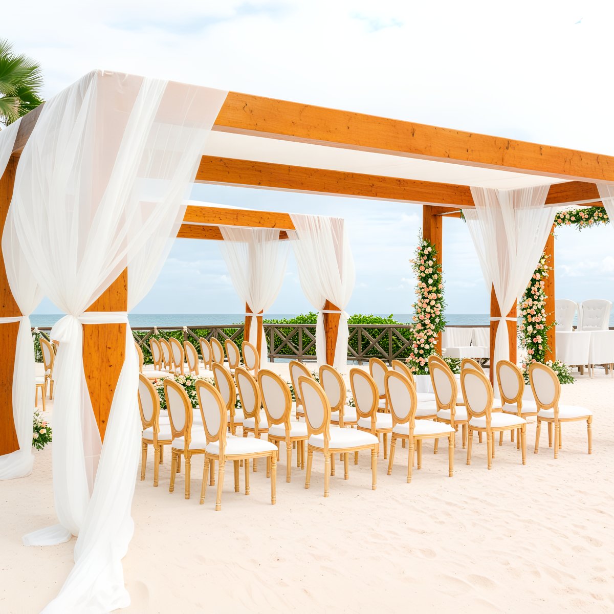 A wooden wedding arch adorned with white drapes stands on the sandy beach, surrounded by chairs arranged for a beautiful destination wedding ceremony.