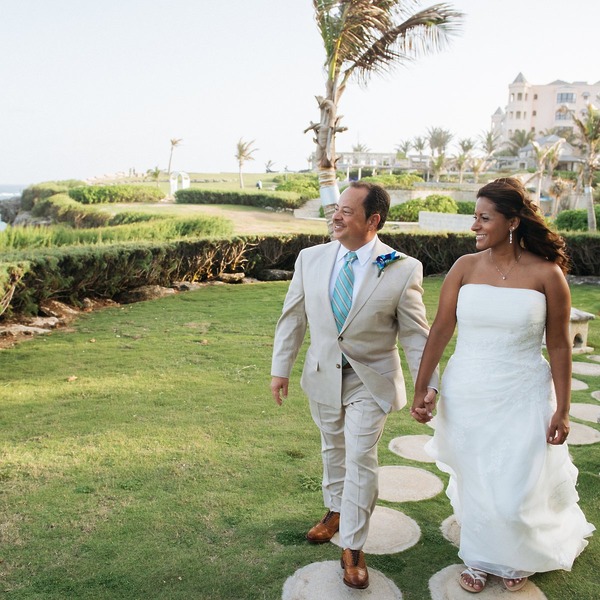 A bride and groom stroll hand in hand on a grassy path at a scenic Barbados destination wedding venue.
