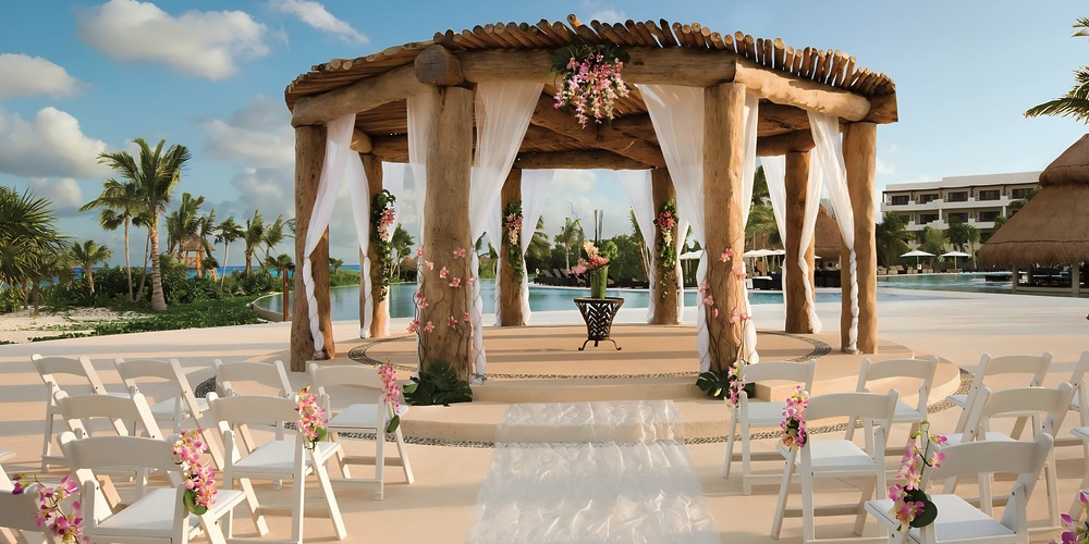 Elegant outdoor destination wedding ceremony setup featuring a wooden gazebo, rows of white chairs, and beautiful floral decorations.