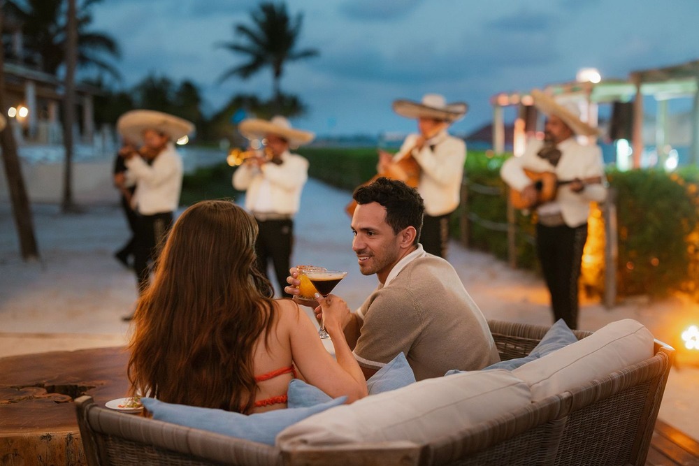 A couple relaxes with drinks on an outdoor couch during their destination wedding honeymoon, while a mariachi band plays nearby.