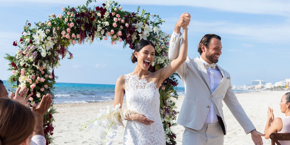 Bride and groom celebrate at a Cancun destination wedding under a floral arch as guests applaud on beach.
