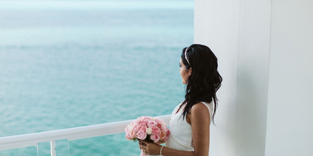 Woman in a white dress stands on a balcony, dreaming of her perfect destination wedding, bouquet of pink flowers in hand—wondering if a destination wedding calculator could make it all possible.