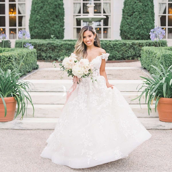 A bride in a stunning white gown stands outdoors by a staircase, offering beautiful inspiration for wedding hair ideas—perfect for your destination wedding look.