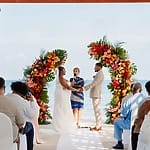 Couple holding hands at a destination wedding by the ocean, surrounded by guests and flowers.
