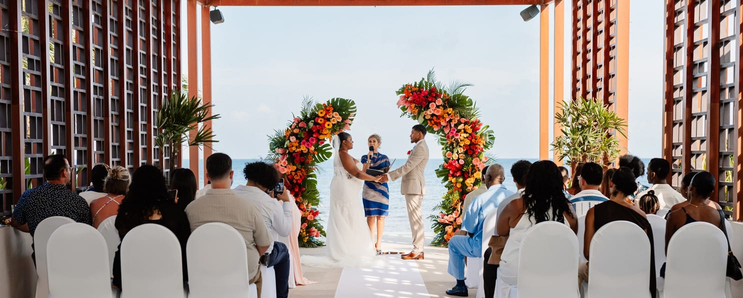 Couple holding hands at a destination wedding by the ocean, surrounded by guests and flowers.