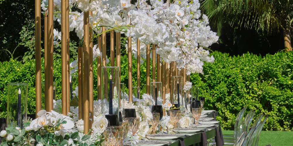 Chic destination wedding table with white floral centerpieces and clear chairs in a lush garden setting.