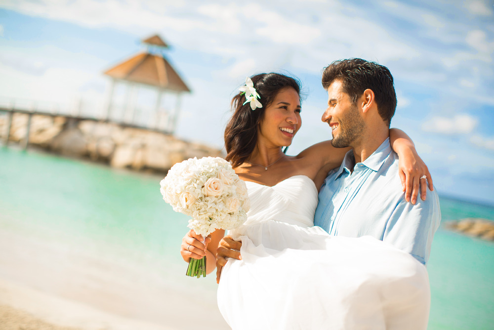 A groom joyfully carries his bride along the beach, both smiling as they imagine their unforgettable destination wedding in Jamaica and consider the cost of their dream wedding.