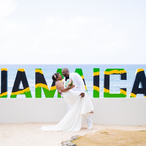 The bride and groom pose beside a vibrant "JAMAICA" sign, capturing the perfect moment for their dream destination wedding in Jamaica.