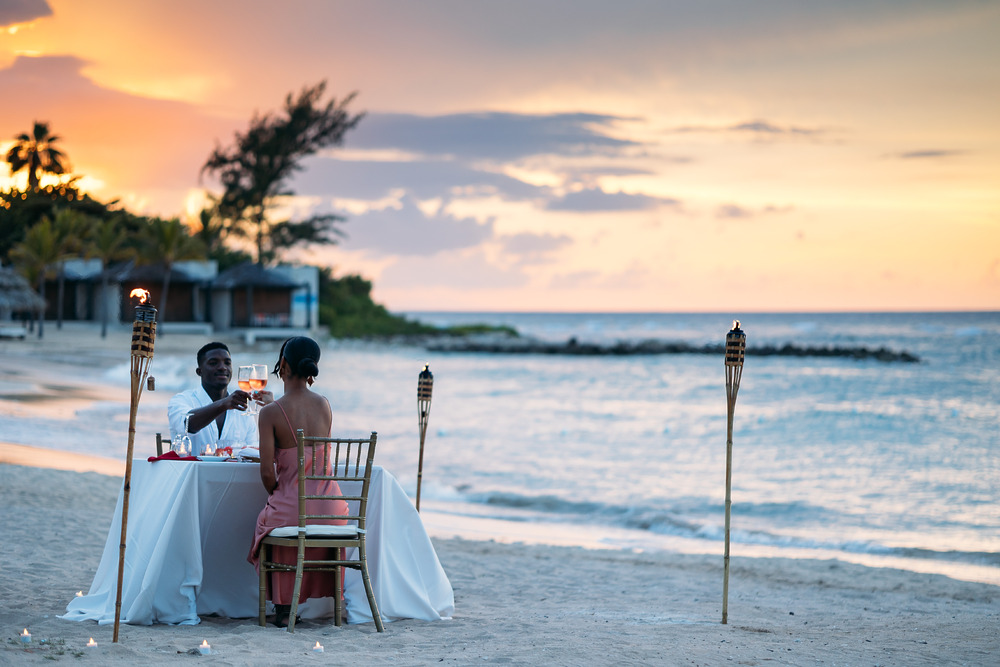 A couple enjoys a romantic sunset dinner on the beach, illuminated by glowing tiki torches—the ideal setting for a destination wedding in Jamaica.