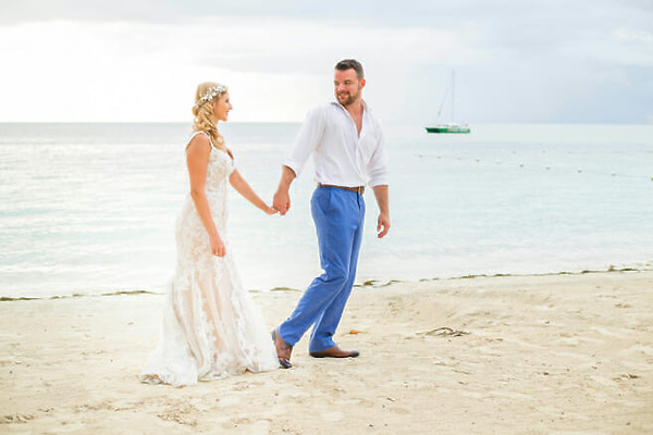 A couple holding hands walks along a beach during their wedding in Jamaica. The woman is wearing a white dress, and the man, dressed in a white shirt and blue pants, beams beside her. A sailboat is visible in the background on the water.