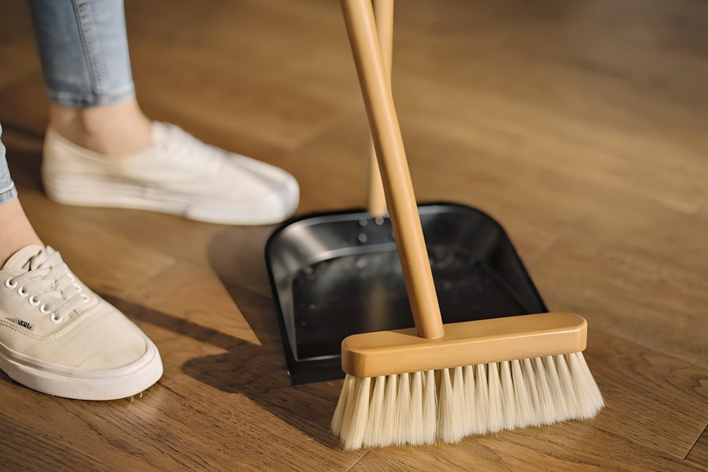 A person sweeps dust into a black dustpan on a wooden floor, tidying up in preparation for a beautiful destination wedding.