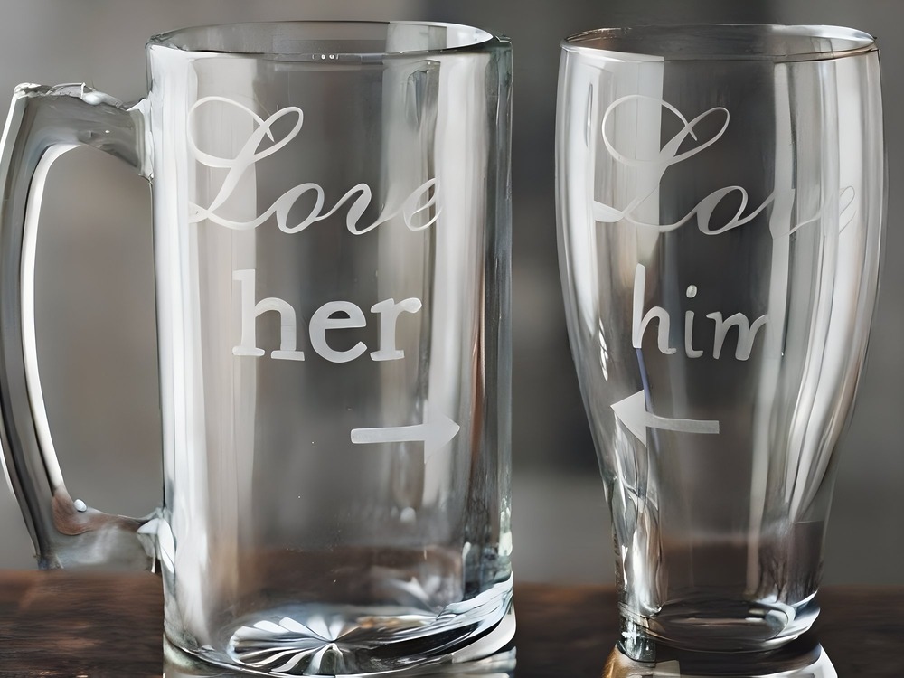 Two glass mugs, each elegantly etched for a wedding or destination wedding—one reads "Love her," the other "Love him," both adorned with playful arrows pointing towards each other.