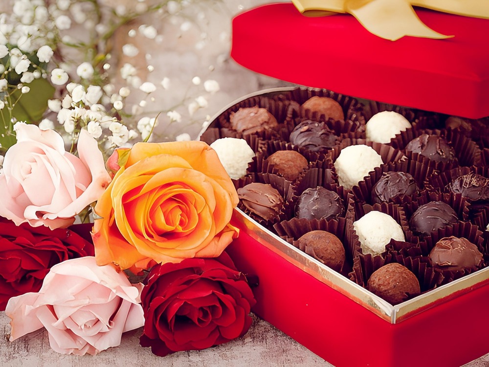 A heart-shaped box of assorted chocolates is elegantly arranged beside delicate roses and baby's breath, creating a romantic display perfect for a wedding or destination wedding celebration.