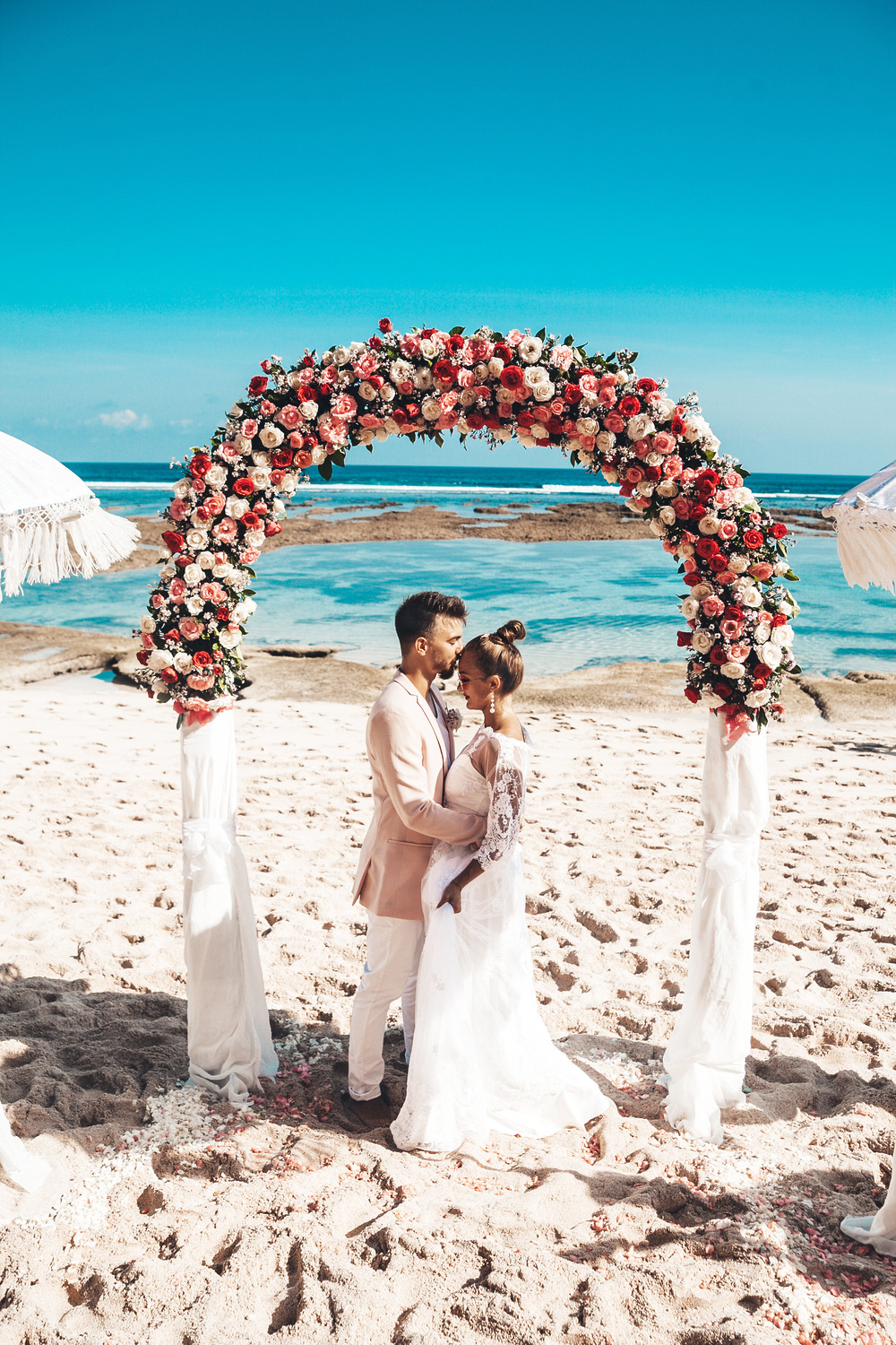 The bride and groom exchange vows under a floral arch on the sandy beach of Tulum, with the blue sky and ocean providing a breathtaking backdrop for their destination wedding.