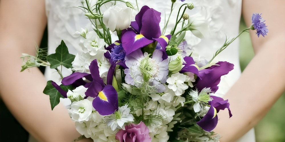 A vibrant spring bouquet of purple, white, and blue flowers is held against a flowing white wedding dress—a stunning accessory for a destination wedding.