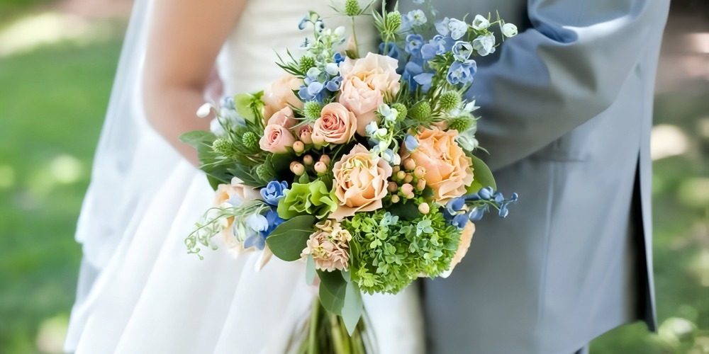 The bride, holding a spring wedding bouquet in soft peach and blue hues, stands beside a person dressed in a gray suit—a perfect moment for their destination wedding.