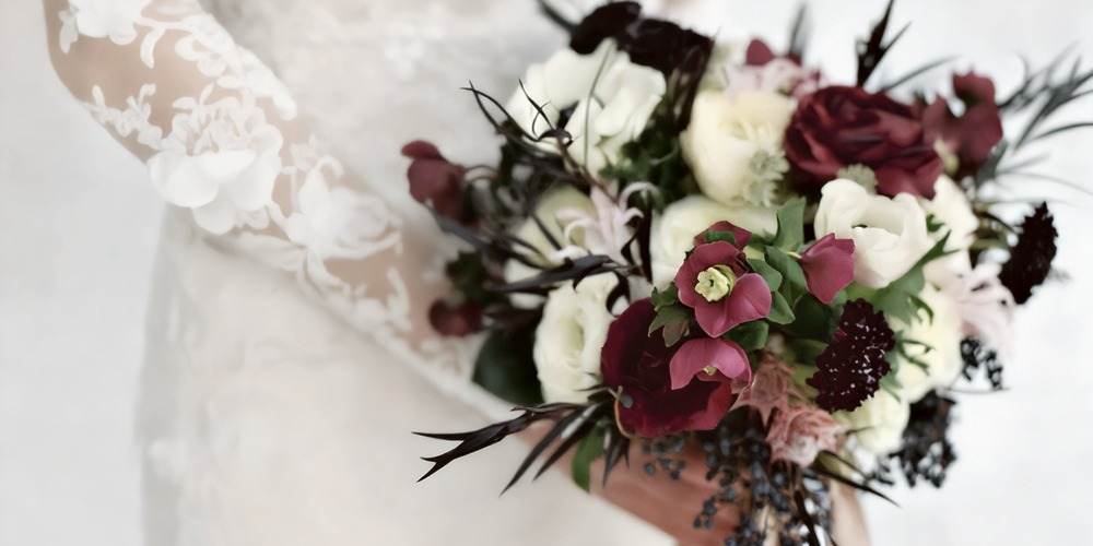 The bride, dressed in lace-sleeved attire, holds a stunning spring wedding bouquet featuring red, white, and deep-hued flowers—perfect for an elegant destination wedding.