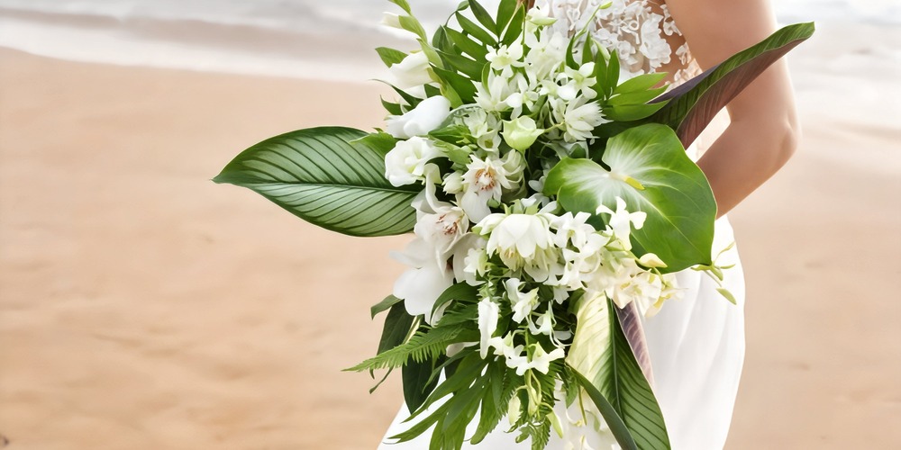 A person holding a lush wedding bouquet with white flowers and green leaves stands on the sandy shore, capturing the essence of a romantic destination wedding by the beach.