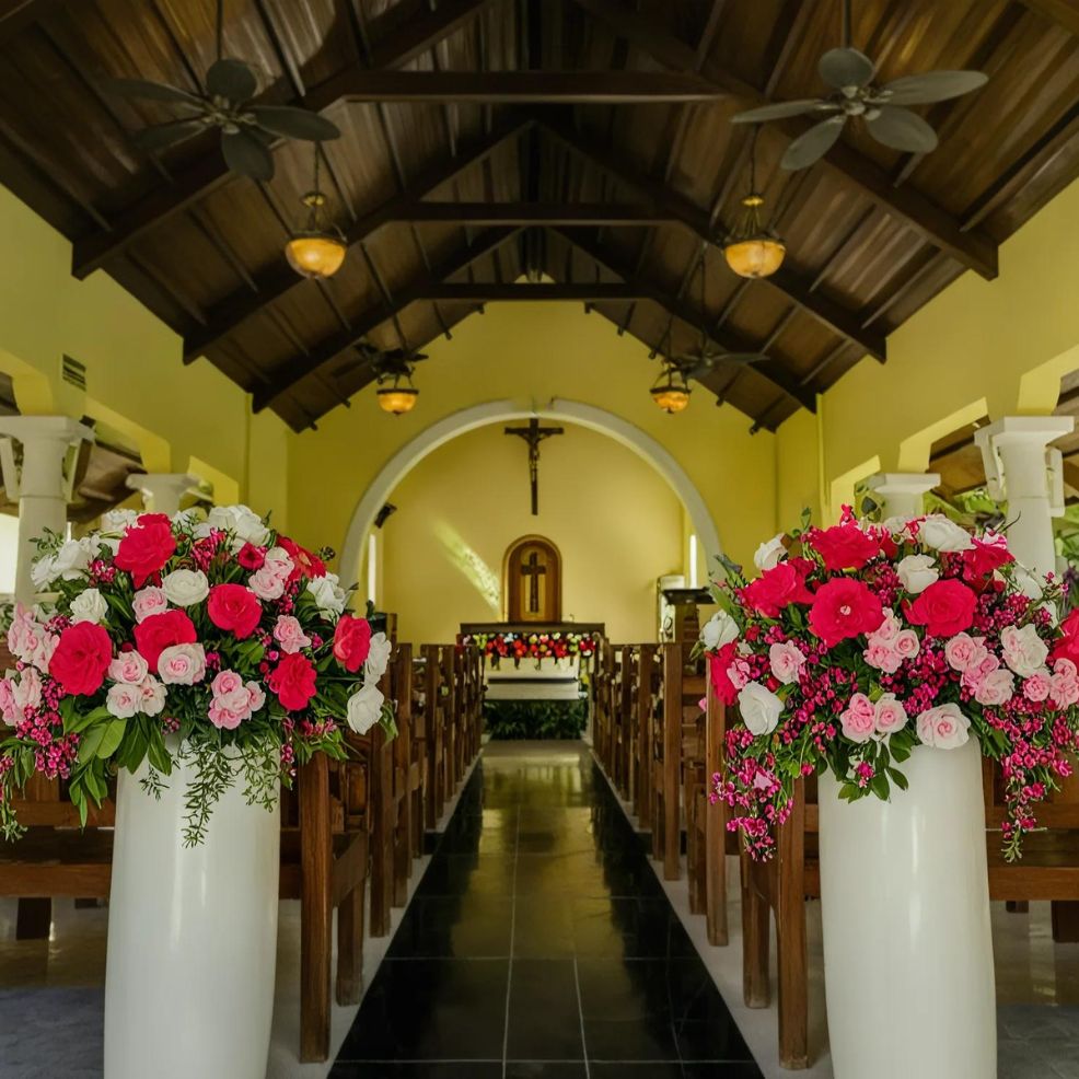 Interior of a church with a central aisle flanked by wooden pews. The space is decorated with arrangements of pink and white flowers, and illuminated by ceiling lights and fans.