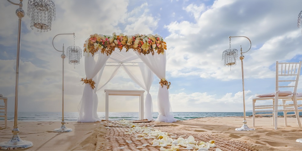 Wedding arch adorned with flowers and drapes at a destination wedding, beach chairs and lanterns nearby.