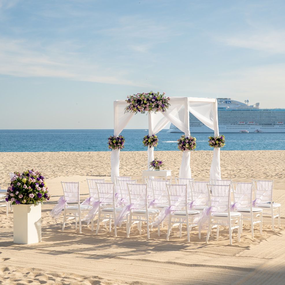 White chairs and a floral arch arranged for a destination wedding on the beach with a cruise ship behind.
