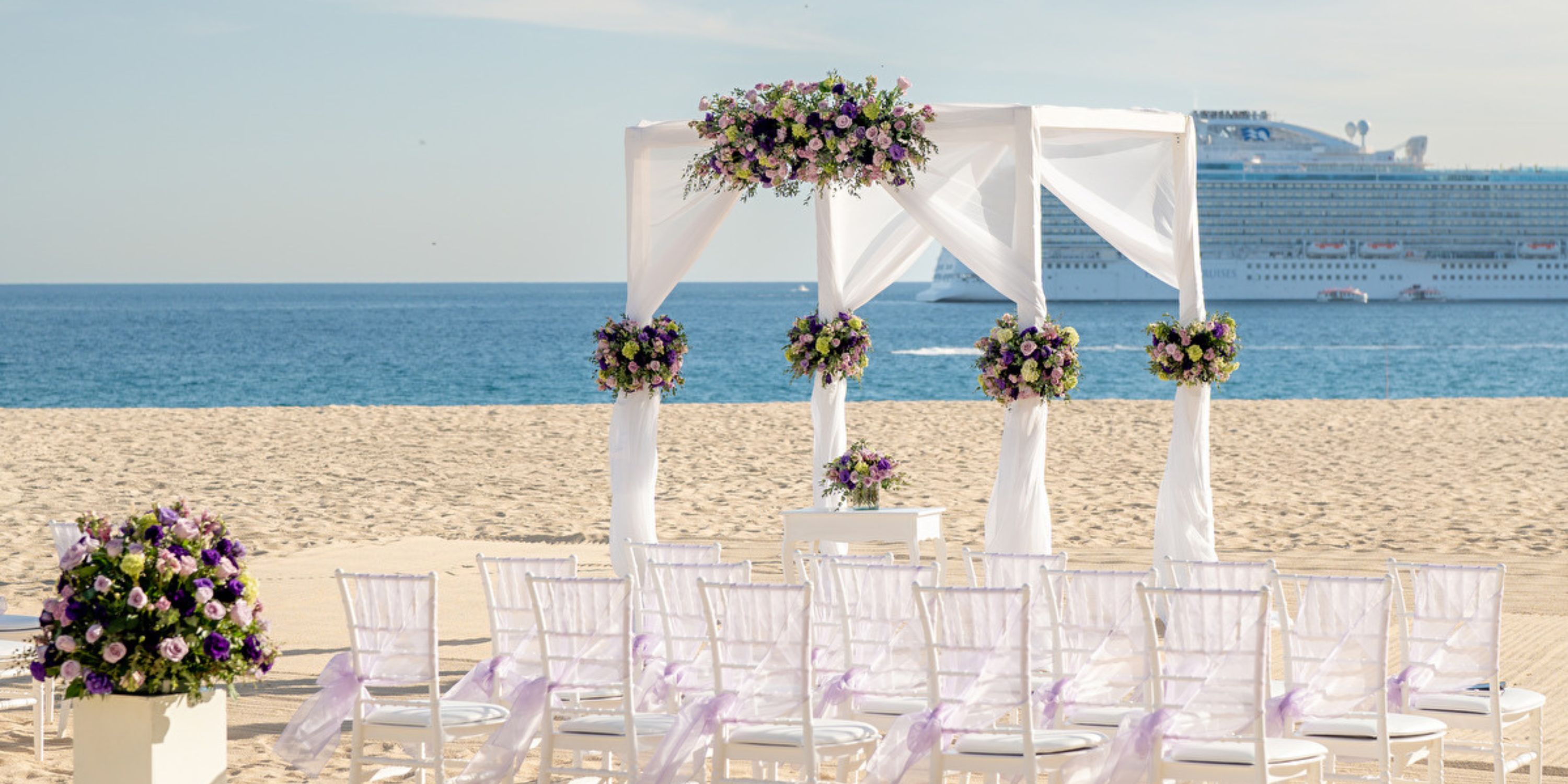 A destination wedding setup on the beach with white chairs, floral accents, and a cruise ship behind.