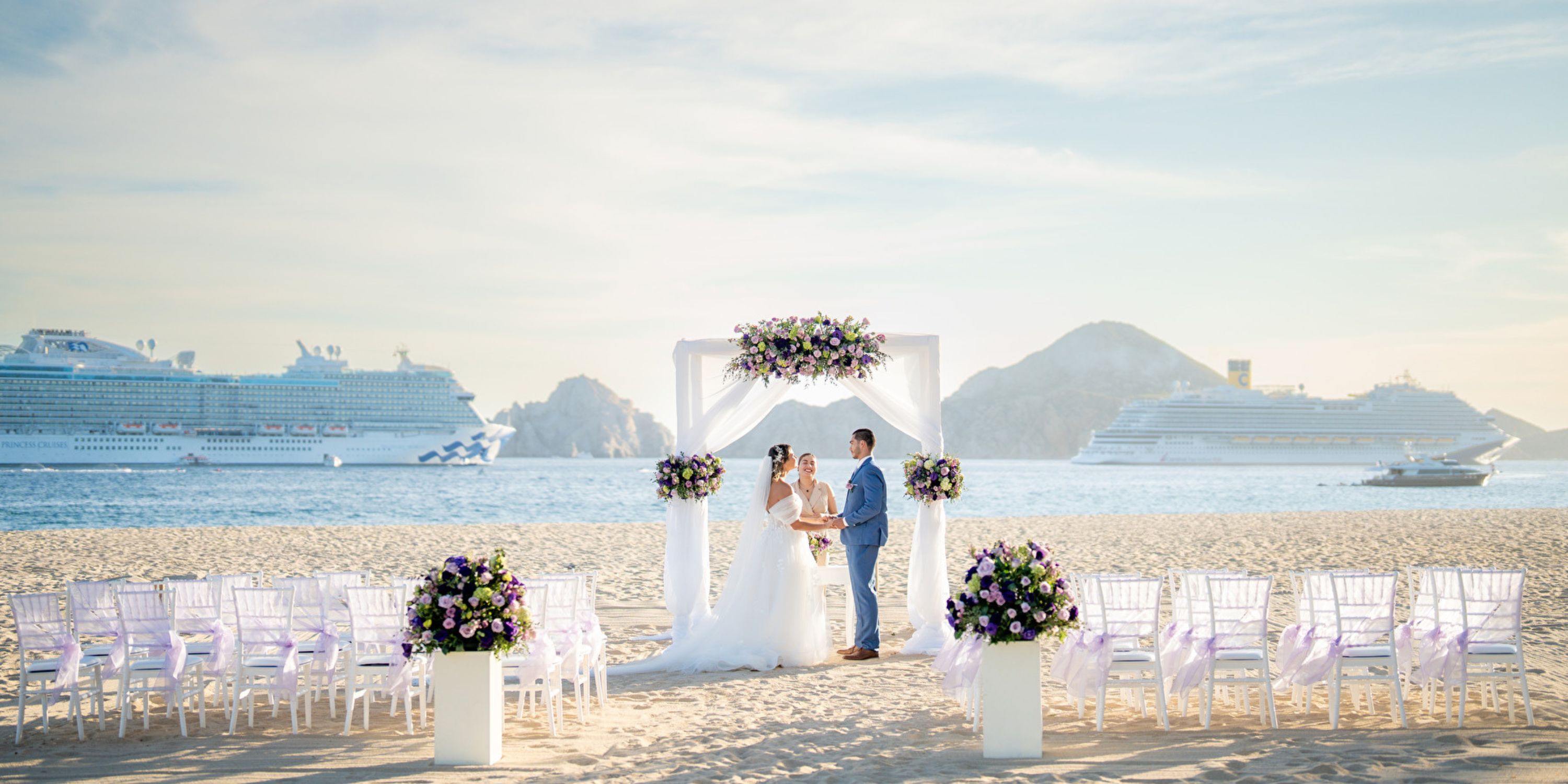 A couple poses beneath a floral arch on a beach during their destination wedding, cruise ships behind them.