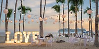 Destination wedding setup on the beach with "LOVE" marquee lights, string lanterns, and elegant tables at sunset.