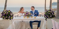 At a destination wedding, the bride and groom sit at a candlelit, purple-flowered table on the beach.