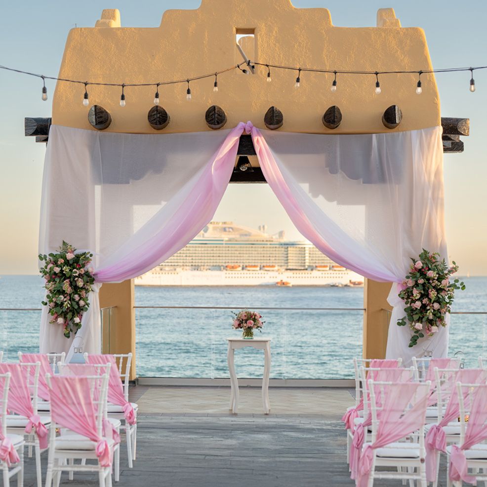 Destination wedding altar adorned with pink drapery by the sea, cruise ship in the background.