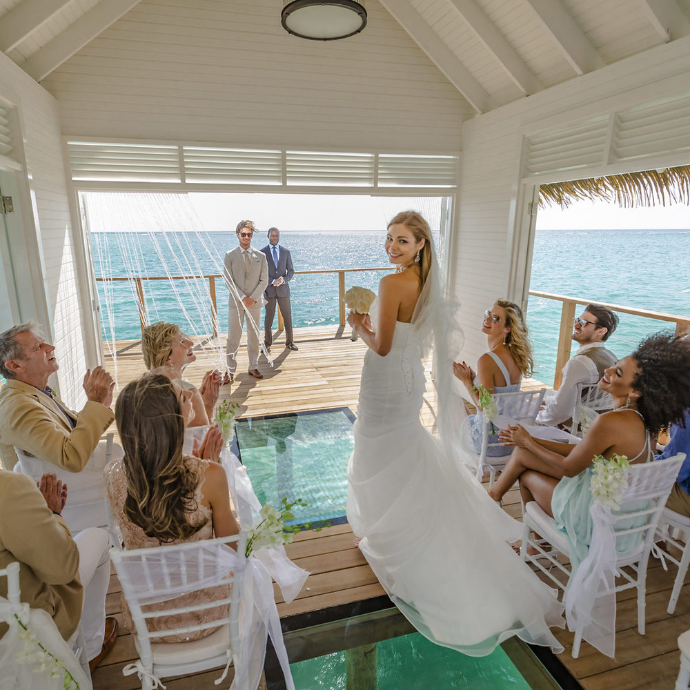 A bride stands and smiles at guests during a beautifully orchestrated wedding ceremony inside a small overwater pavilion with clear ocean views, embracing the charm of free weddings.