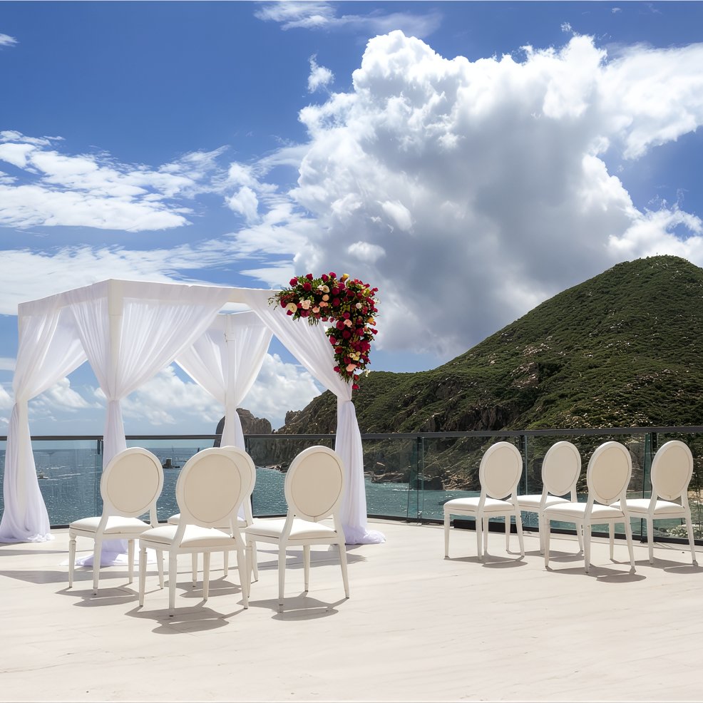 White chairs and a floral canopy set for a destination wedding overlook the ocean and green hills below.