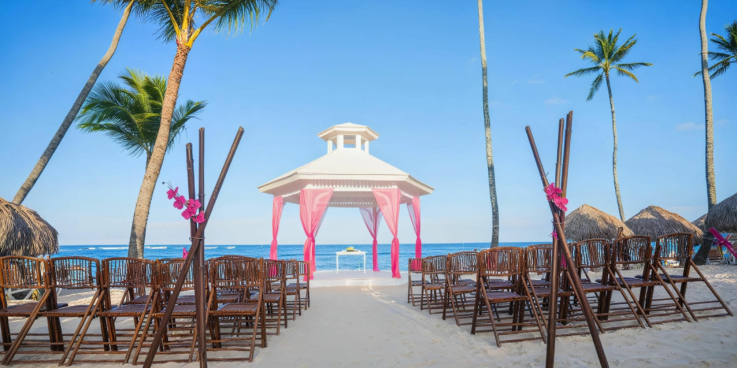 An outdoor beach wedding setup featuring the Majestic Colonial includes a white gazebo adorned with pink fabric, surrounded by wooden chairs and palm trees, overlooking the ocean under a clear blue sky. This scene captures pure romance and tropical elegance.