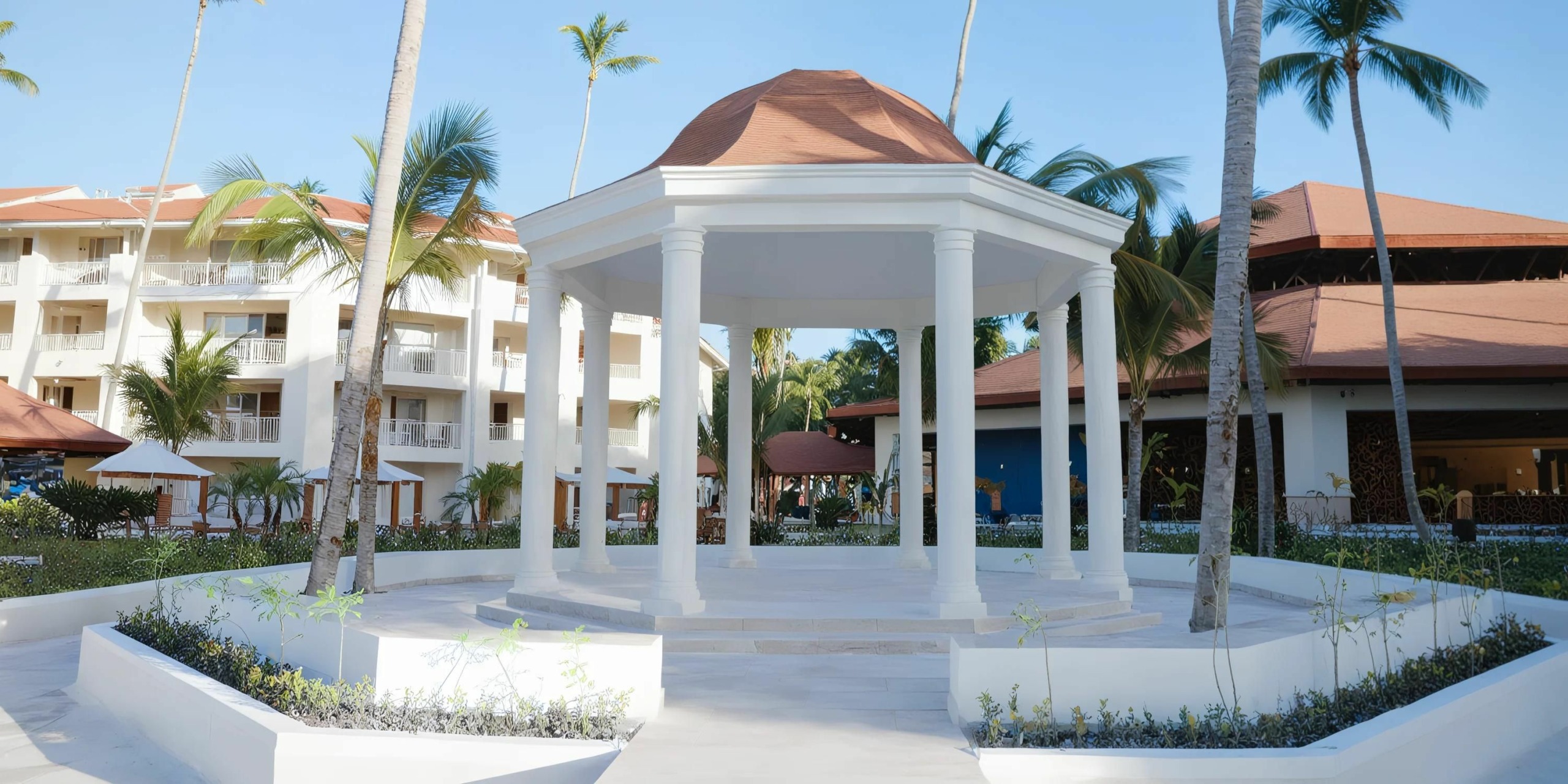 A white gazebo with a brown roof is surrounded by palm trees in an outdoor area, near buildings with red-tiled roofs. The clear sky provides the perfect backdrop for weddings at Majestic Mirage.