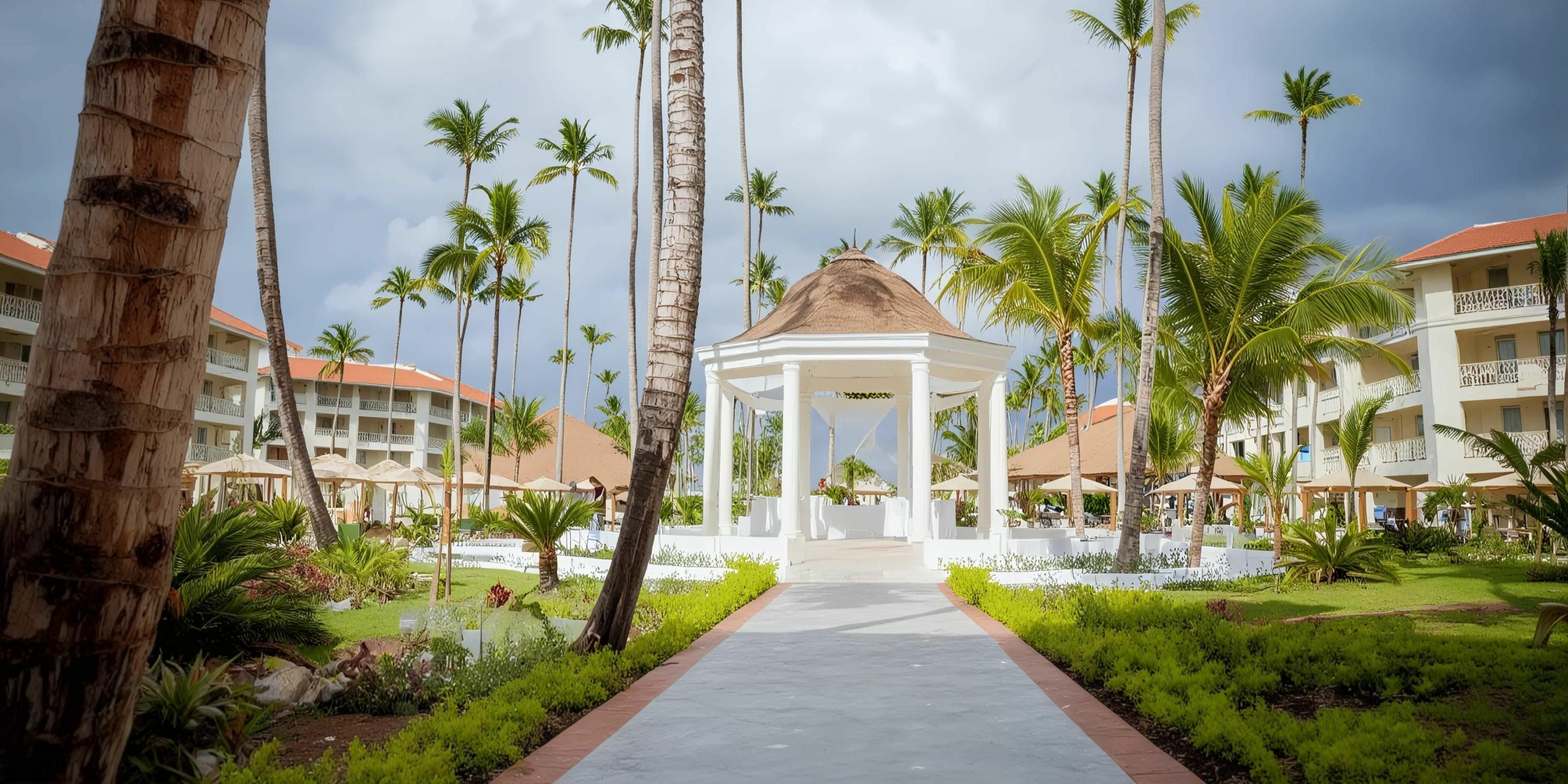 A white gazebo with a thatched roof stands in a tropical garden, surrounded by palm trees and resort buildings, perfect for weddings at Majestic Mirage.