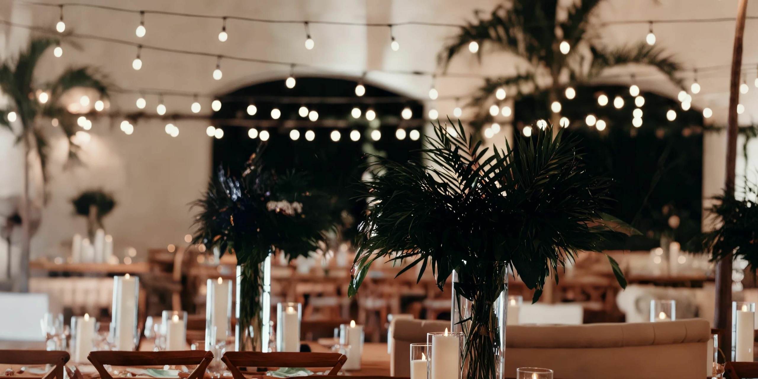 A decorated event space with string lights and greenery centerpieces on tables.