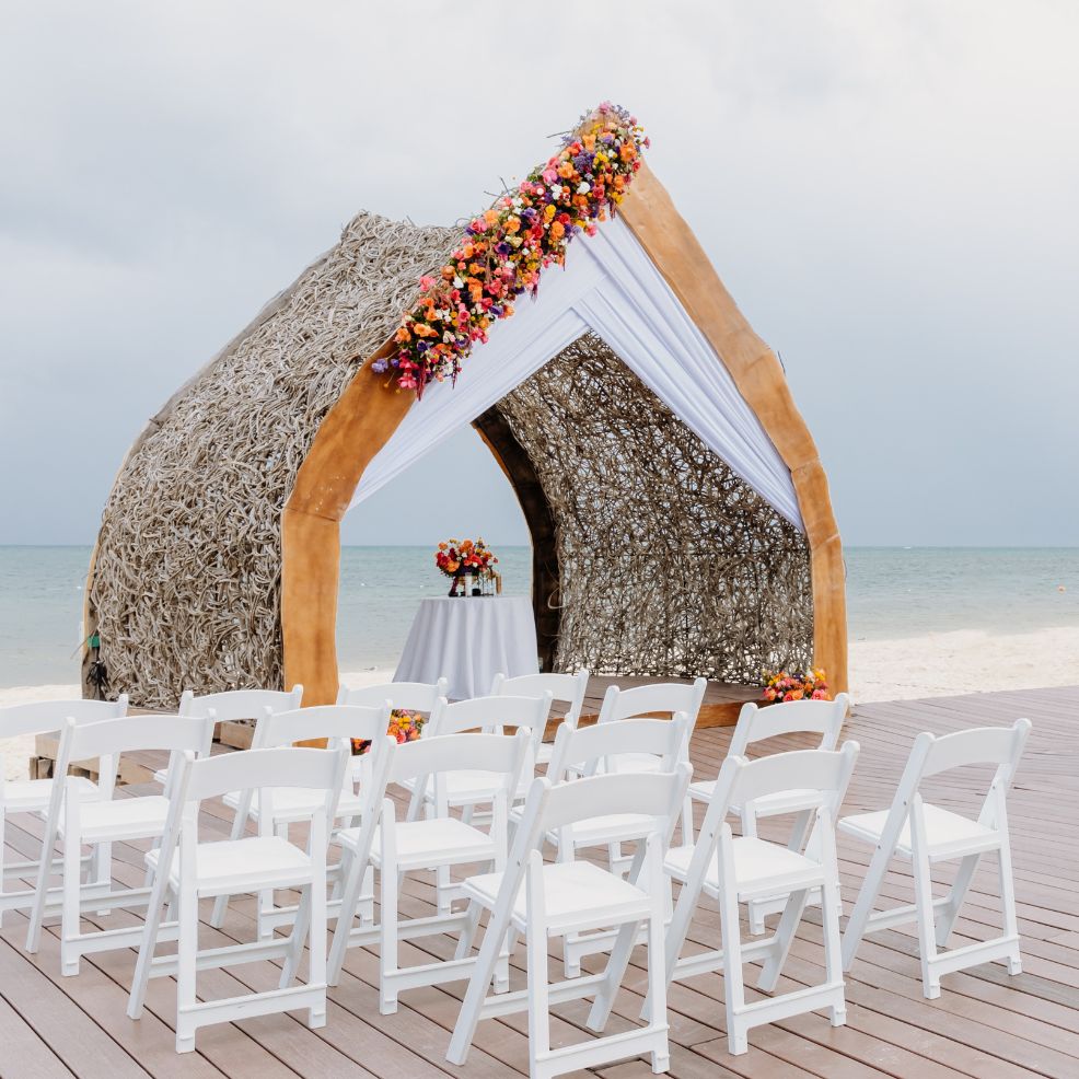 Destination wedding setup on the beach with rows of white chairs and a floral arch overlooking the ocean.