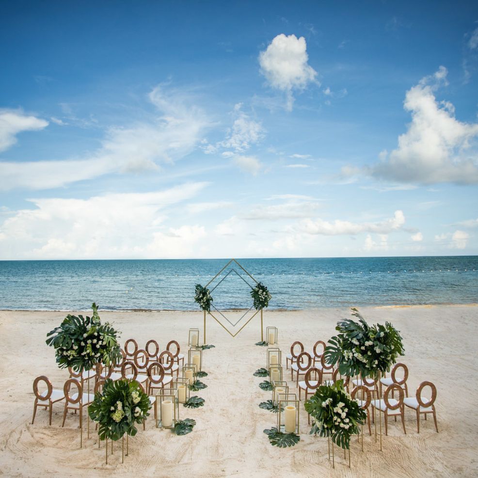 Destination wedding setup on the beach with chairs, floral decor, and a geometric arch overlooking the ocean.