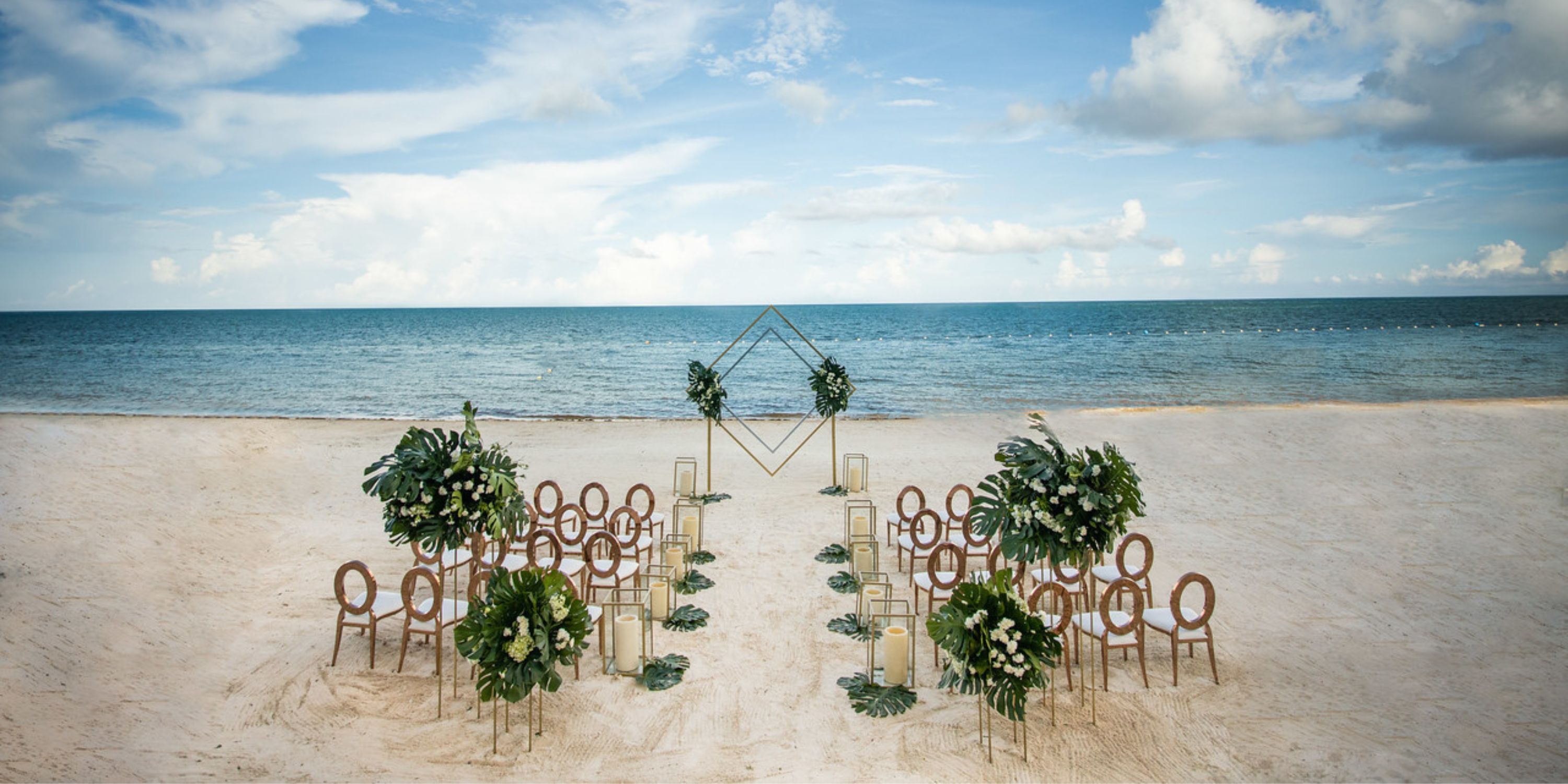 Destination wedding setup on the beach with chairs and floral décor arranged toward the ocean under blue sky.