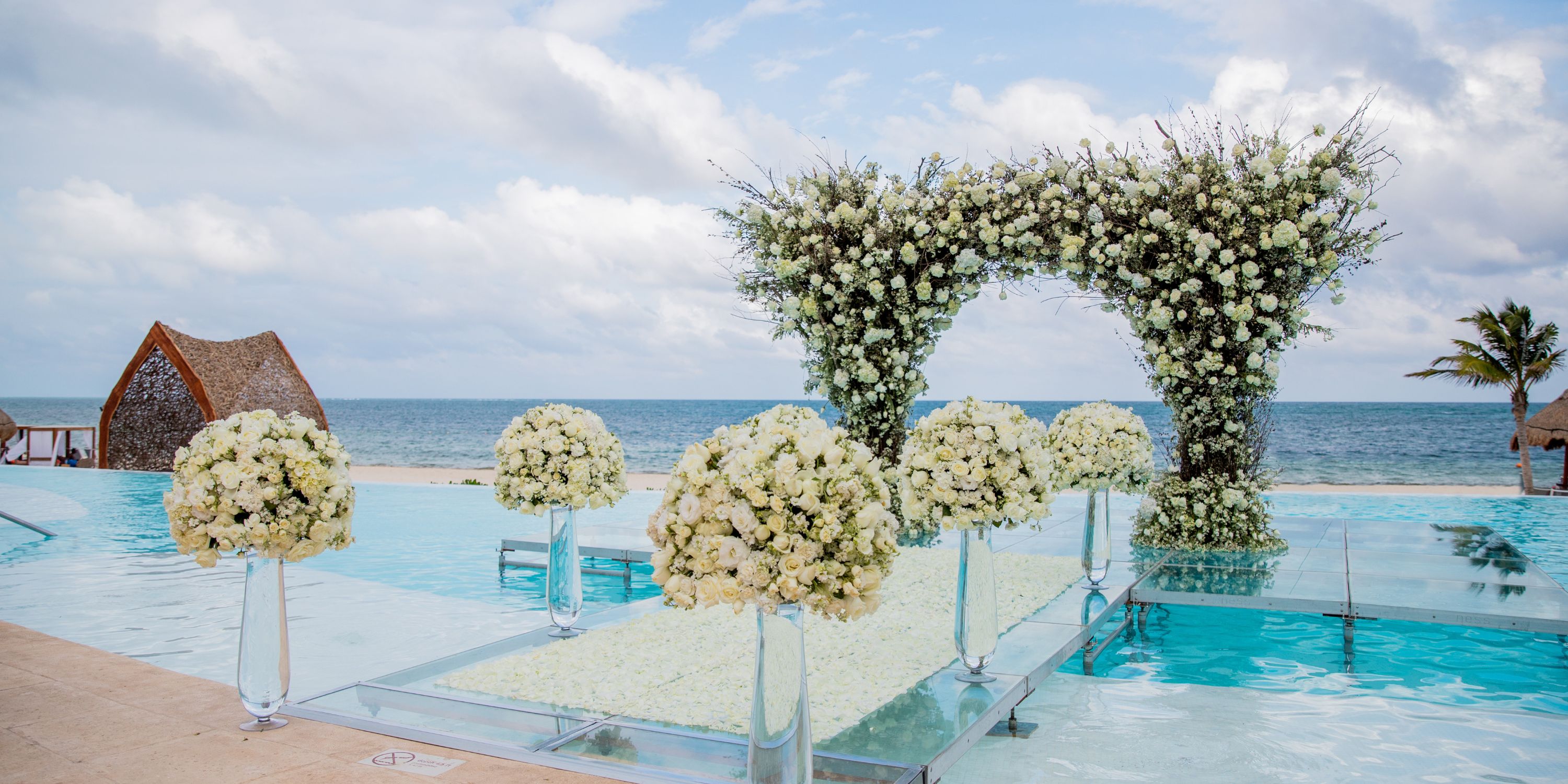 Destination wedding arch and floral arrangements displayed on a platform above a pool by the ocean.
