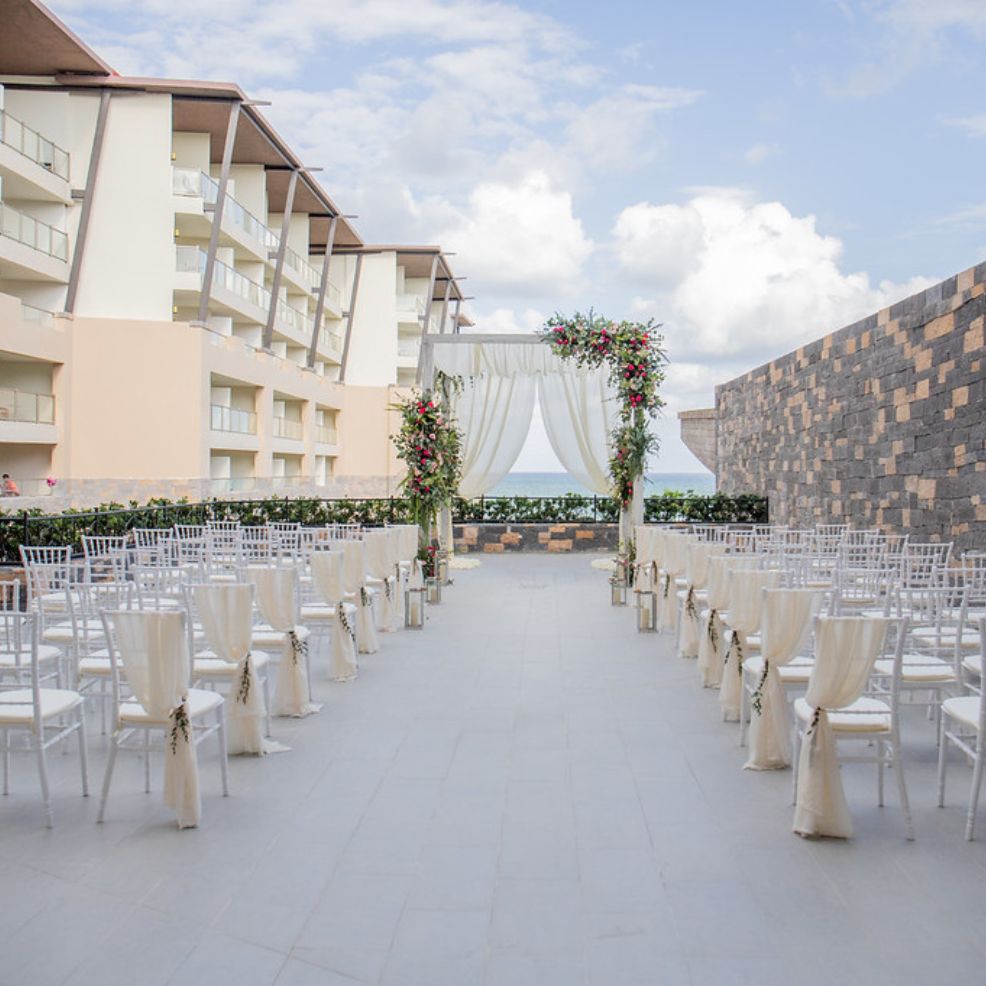 Destination wedding ceremony featuring white chairs and a floral arch set before a hotel backdrop.